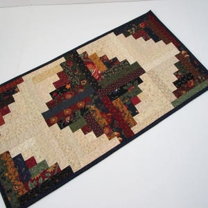 May include: A rectangular quilted table runner with a log cabin design. The center features a cream-colored fabric surrounded by strips of fabric in various colors, including red, green, and navy. The runner is bordered with a dark blue fabric.