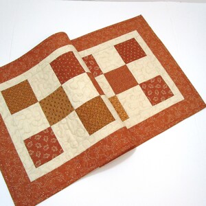 May include: A brown and cream table runner with a patchwork design. The runner is made of different fabrics with various patterns, including small dots and floral designs. The runner has a brown border with a subtle floral pattern.