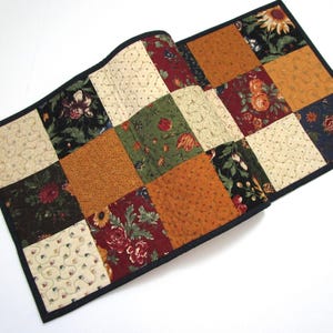 May include: A patchwork table runner with a black border. The squares feature floral and patterned fabrics in shades of burgundy, gold, green, and cream. The runner is approximately 12 inches wide and 24 inches long.