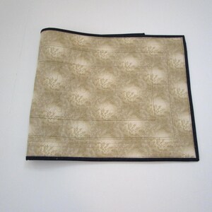 May include: A beige placemat with a floral pattern and a black border. The placemat is rectangular and appears to be made of fabric. The floral pattern is a lighter shade of beige.