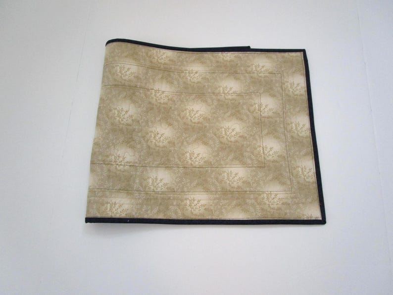May include: A beige placemat with a floral pattern and a black border. The placemat is rectangular and appears to be made of fabric. The floral pattern is a lighter shade of beige.