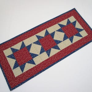 May include: A rectangular quilted table runner with a patriotic theme. The runner features a red border with a small white dot pattern, a central design of red squares and blue stars on a beige background, and a blue border.