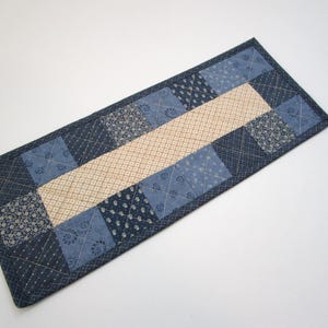 May include: A rectangular quilted table runner featuring a central strip of cream fabric with a small dark blue grid pattern, bordered by alternating squares of various blue fabrics with floral, dotted, and geometric patterns, all stitched with a cross-hatch design.