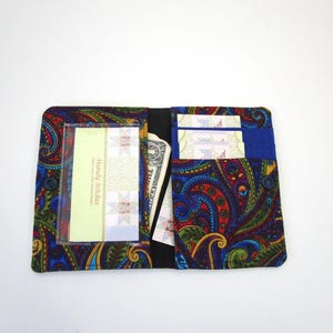 May include: A fabric wallet featuring a vibrant multi-colored paisley pattern in shades of blue, green, red, and yellow. The interior includes a clear plastic ID window, multiple card slots, and a pocket containing paper currency.