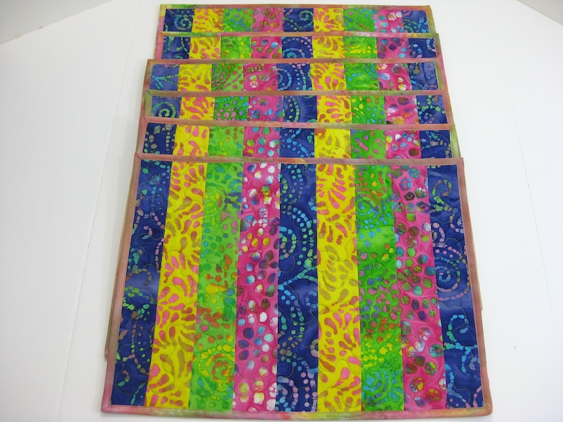 May include: Set of six colorful placemats with a striped pattern. The placemats are made of fabric and have a batik design. The colors are blue, green, yellow, and pink.