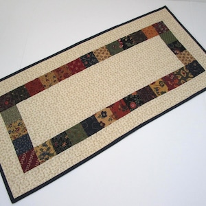 May include: Rectangular quilted table runner with a cream-colored center and a border of colorful patchwork squares. The border is framed by a thin black edge. The patchwork includes floral and patterned fabrics in various colors.