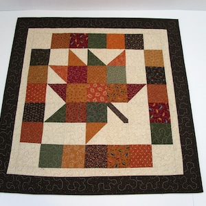 May include: A square quilt with a leaf design in the center. The quilt features a variety of fall colors, including orange, brown, green, and red. The leaf is surrounded by a cream-colored background and a dark brown border.