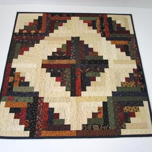 May include: A rectangular quilt featuring a log cabin pattern with a central square block. The design incorporates various fabric strips in shades of cream, dark red, forest green, navy blue, and mustard yellow, finished with a dark blue border.