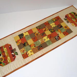 May include: A rectangular table runner featuring a central panel of patchwork squares in shades of orange, yellow, green, and brown. Two apple-shaped appliqués with vertical fabric strips in similar autumn colors are positioned at each end of the runner.