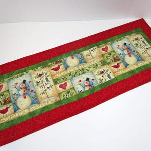 May include: A rectangular fabric table runner featuring a red border with swirling quilted patterns and a green inner border. The center panel displays a repeating winter scene with snowmen, red birds, hearts, and the text Winter Wonders.