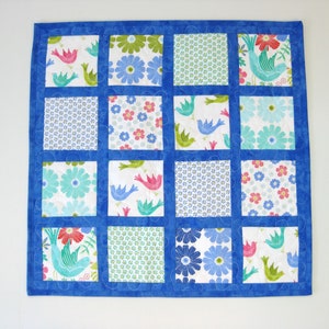 May include: A blue and white patchwork quilt with a variety of floral and bird patterns. The quilt is made up of 16 squares, each with a different design. The quilt is edged in blue fabric.