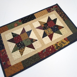 May include: A rectangular quilted table runner featuring two central star patterns in shades of burgundy, navy, olive green, and tan. The stars are set against a cream background with swirling stitch details, surrounded by a border of floral fabric.