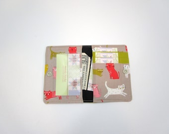 May include: Open fabric wallet with a cat pattern in red, white, and green on a tan background. The wallet is open, revealing card slots and a cash compartment with a dollar bill. A button closure is visible.