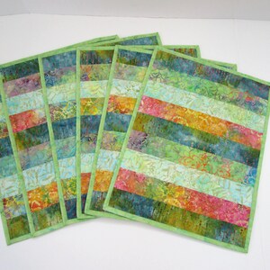 May include: A set of five rectangular fabric placemats featuring horizontal stripes in various batik patterns. The colors include shades of green, blue, yellow, orange, and pink. Each placemat is finished with a solid light green fabric border.