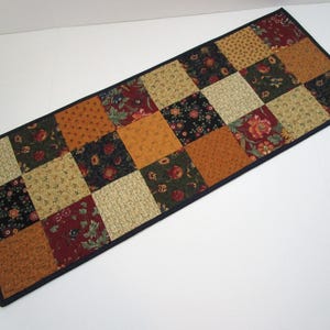 May include: A rectangular patchwork table runner featuring a grid of squares in various autumnal colors. The squares display floral and geometric patterns in shades of gold, red, and green, with a dark blue border.