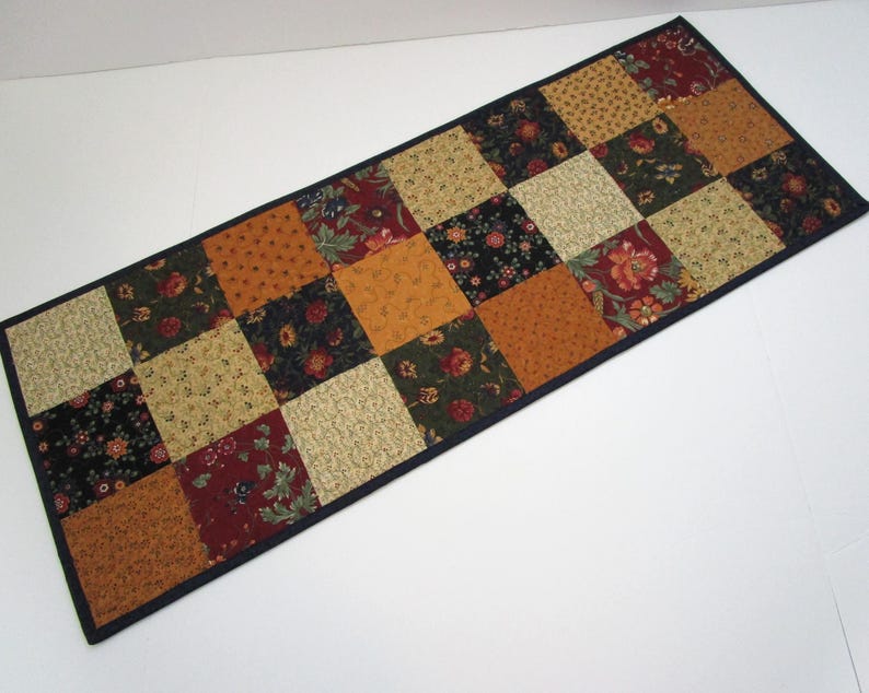 May include: A rectangular patchwork table runner featuring a grid of squares in various autumnal colors. The squares display floral and geometric patterns in shades of gold, red, and green, with a dark blue border.