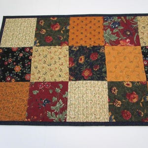May include: A rectangular quilted table runner with a patchwork design. The squares feature floral patterns in shades of orange, red, green, and cream, set against a dark blue border. The runner is likely handmade.