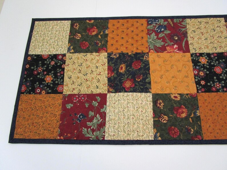 May include: A rectangular quilted table runner with a patchwork design. The squares feature floral patterns in shades of orange, red, green, and cream, set against a dark blue border. The runner is likely handmade.