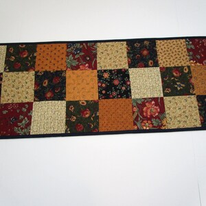 May include: A rectangular patchwork table runner featuring a variety of patterned squares in shades of red, gold, green, and tan. The squares are arranged in a grid pattern and bordered by a dark blue fabric.