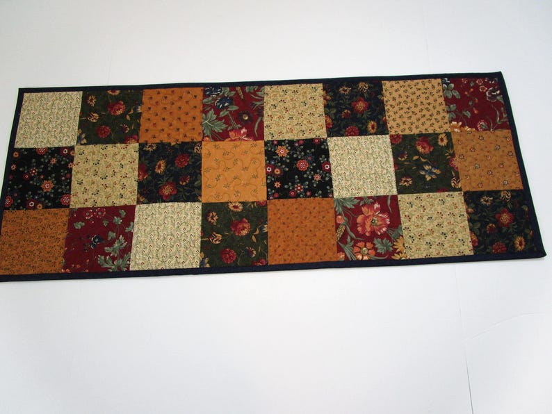 May include: A rectangular patchwork table runner featuring a variety of patterned squares in shades of red, gold, green, and tan. The squares are arranged in a grid pattern and bordered by a dark blue fabric.