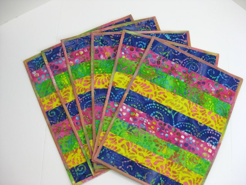 May include: Set of six colorful placemats with a striped pattern. The placemats are made of fabric with a batik design in shades of pink, yellow, green, and blue.