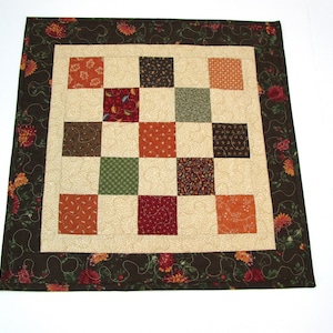 May include: A square patchwork quilt with a brown floral border. The quilt is made up of 16 smaller squares in various shades of brown, green, orange, and red.