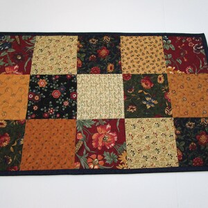 May include: A rectangular patchwork table runner with a dark blue border. The runner features a variety of floral and patterned fabrics in shades of red, gold, green, and black. The design is a grid of squares.