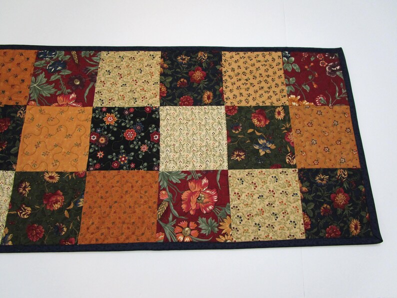 May include: A rectangular patchwork table runner with a dark blue border. The runner features a variety of floral and patterned fabrics in shades of red, gold, green, and black. The design is a grid of squares.