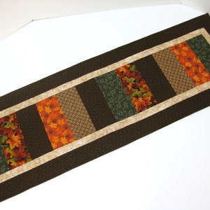 May include: A brown table runner with a beige border and a patchwork design of green, orange, and brown fabric. The fabric features a leaf pattern.