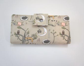 May include: A beige wallet with a whimsical forest pattern featuring sleeping animals, moons, and trees. The wallet has a snap closure and a rectangular shape. The design includes shades of brown, gray, and white.