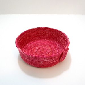 May include: A pink woven rope bowl with a white swirl pattern. The bowl is round and has a slight lip around the edge.