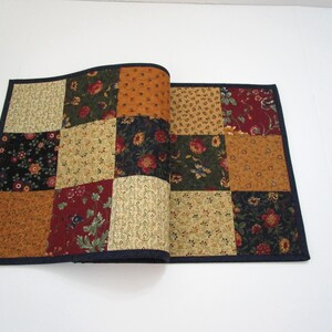 May include: A patchwork table runner with a dark blue border. The squares feature floral and geometric patterns in shades of gold, red, green, and black. The runner is folded open, displaying the interior fabric.