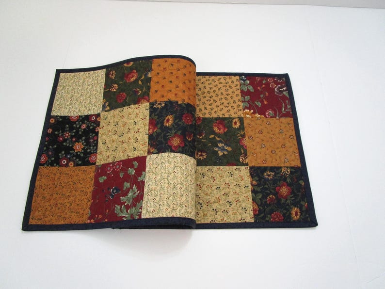 May include: A patchwork table runner with a dark blue border. The squares feature floral and geometric patterns in shades of gold, red, green, and black. The runner is folded open, displaying the interior fabric.