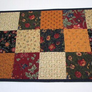 May include: A patchwork table runner featuring a variety of floral patterns in shades of red, orange, green, and cream. The squares are bordered by a dark blue trim. The runner is approximately 36 inches long and 18 inches wide.
