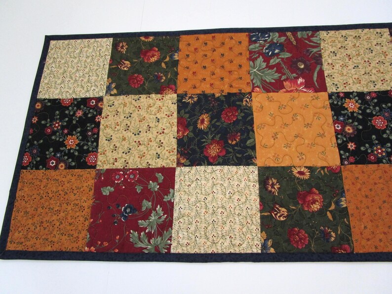 May include: A patchwork table runner featuring a variety of floral patterns in shades of red, orange, green, and cream. The squares are bordered by a dark blue trim. The runner is approximately 36 inches long and 18 inches wide.