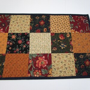 May include: A patchwork table runner featuring a variety of floral patterns in shades of red, gold, green, and black. The runner is bordered with a dark blue fabric. The design is a mix of traditional and contemporary styles.