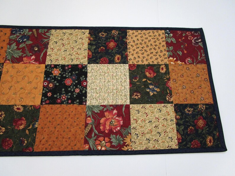 May include: A patchwork table runner featuring a variety of floral patterns in shades of red, gold, green, and black. The runner is bordered with a dark blue fabric. The design is a mix of traditional and contemporary styles.