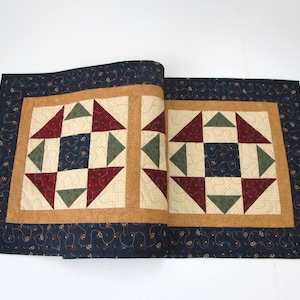 May include: A rectangular quilted table runner featuring two square blocks with geometric patterns in dark blue, cream, maroon, and olive green. The border is a dark blue fabric with a swirling vine and small flower print, framed by a solid tan border.