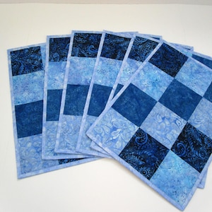 May include: A set of six blue fabric placemats. Each placemat features a patchwork design with squares and stripes in various shades of blue, including a darker paisley pattern. The placemats have a light blue border.