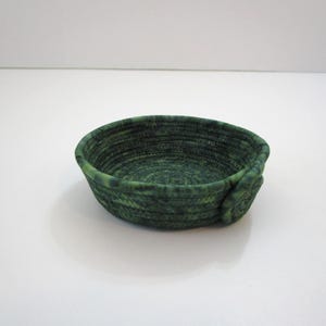 May include: A small, round, green bowl with a textured, woven appearance. The bowl is a deep shade of green with lighter green accents, and has a small, decorative detail on the side. The bowl is likely handmade and suitable for small items.