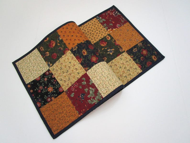 May include: A patchwork quilt with a dark blue border. The quilt is made of squares in various colors, including gold, orange, red, and green, with floral patterns. The quilt is partially folded over.