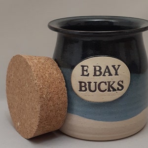 May include: A ceramic jar with a cork lid. The jar is decorated with a blue and white glaze and has the words "E Bay Bucks" written on it.