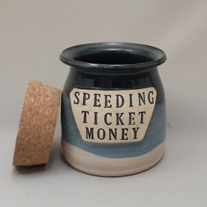 May include: A blue and white ceramic jar with a cork lid. The jar has a white label with the text "SPEEDING TICKET MONEY".