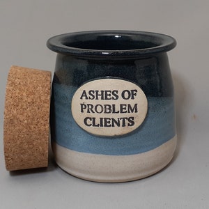 May include: A blue and white ceramic jar with a cork lid. The jar has a white oval label with the text "Ashes of Problem Clients".
