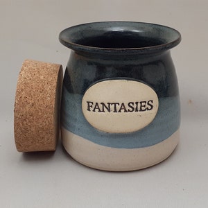 Puede incluir: Un tarro de cerámica azul y blanco con tapa de corcho. El tarro tiene una etiqueta ovalada blanca con la palabra "Fantasies" escrita en negro.