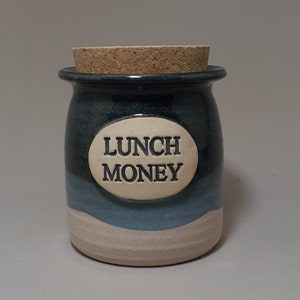 Puede incluir: Un tarro de cerámica azul y blanco con tapa de corcho. El tarro tiene un óvalo blanco con el texto "LUNCH MONEY" en negro.