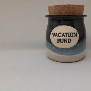 May include: A ceramic jar with a cork lid, painted in shades of blue and black. The jar has a white oval label with the text "VACATION FUND" in black lettering.