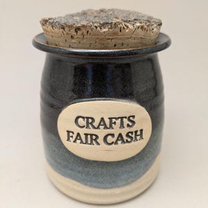 May include: A handmade ceramic jar with a cork lid. The jar is dark blue with a lighter blue and cream-colored base. The text "CRAFTS FAIR CASH" is on an oval label on the front. This jar is perfect for storing small items.