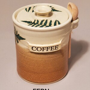 May include: A white ceramic coffee jar with a brown glaze and a green fern design. The jar has a lid with a wooden spoon attached. The jar is labeled "COFFEE" and the word "FERN" is printed below the jar.