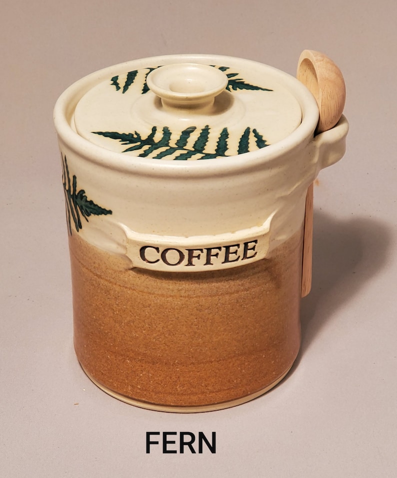 May include: A white ceramic coffee jar with a brown glaze and a green fern design. The jar has a lid with a wooden spoon attached. The jar is labeled "COFFEE" and the word "FERN" is printed below the jar.